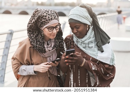 Two friends in muslim clothing discussing photos online on mobile phone during their walk in the city