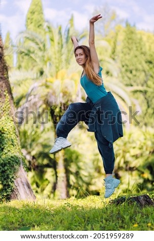 Girl in turquoise hip-hop clothes frozen in a jump in a position of hands up and raise a knee in a summer green park on a sunny day