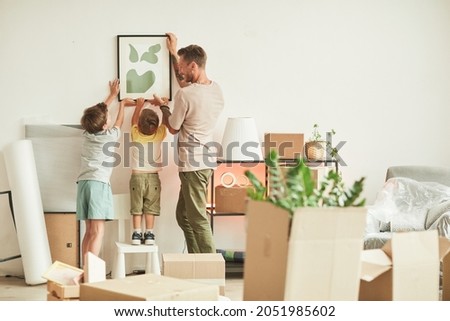Full length portrait of happy father with two sons hanging pictures on wall while moving in to new home, copy space