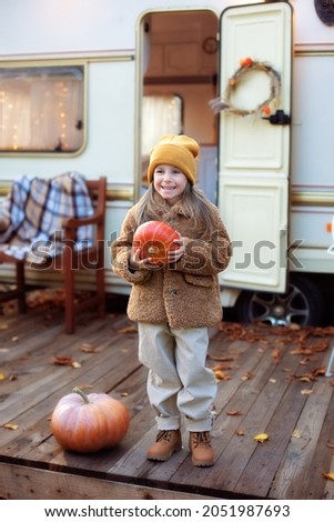 Happy Halloween. Smiling girl standing on the porch with a pumpkin in her hands. Trick-or-treat. Happy little girl playing near camping at RV home in the garden. Child in cozy campsite fall backyard. 