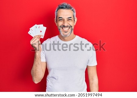 Handsome middle age man with grey hair holding poker cards looking positive and happy standing and smiling with a confident smile showing teeth 