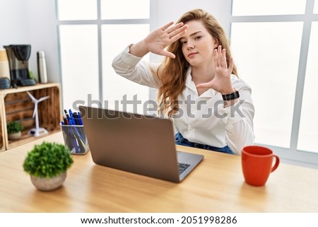 Young caucasian woman working at the office using computer laptop doing frame using hands palms and fingers, camera perspective 