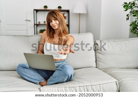 Hispanic woman sitting on the sofa at home using laptop smiling cheerful offering palm hand giving assistance and acceptance. 