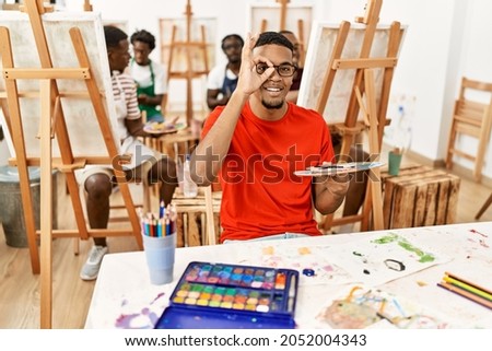 Young african man at art studio smiling happy doing ok sign with hand on eye looking through fingers 