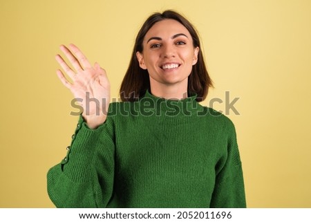 Young woman in green warm sweater happy smiling waving hand to camera welcoming