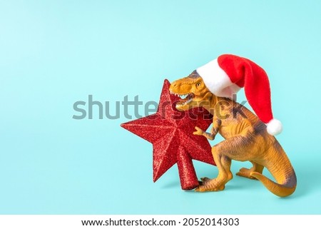 Dinosaur Rex in red Santa Claus hat holds star in its paws on blue background New Years Eve or Christmas Eve Art holiday card Creative idea for Merry xmas concept 