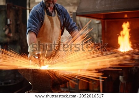 Blacksmith in apron forging the iron with hammer while working in the workshop Royalty-Free Stock Photo #2052016517