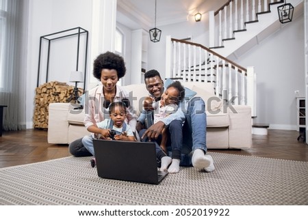 African american family of four sitting together on floor and watching cartoon on laptop. Young parents with two daughters using technology during free time at home.