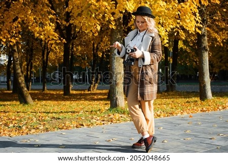 Young hipster woman in hat and boho clothes with retro film camera standing in autumn park outdoors. Girl tourist photographer, hobby.