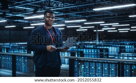Portrait of a Data Center Engineer Using Laptop Computer. Server Room Specialist Facility with African American Male System Administrator Working with Data Protection Network for Cyber Security. Royalty-Free Stock Photo #2052055832