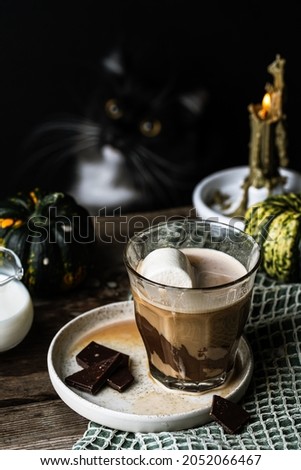 
Halloween coffee. Glass of Mocha latte with marshmallow , chocolate and spices on old wooden table with pumpkins and lit candle and black cat on dark background.