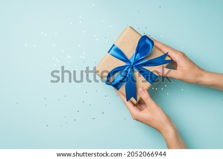 First person top view photo of woman's hands giving craft paper giftbox with blue satin ribbon bow over shiny sequins on isolated pastel blue background with blank space
