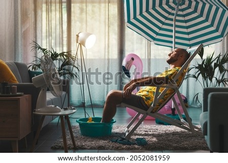 Bored man spending summer vacations at home and pretending he is on the beach, he is sunbathing in his living room and resting on a deckchair Royalty-Free Stock Photo #2052081959