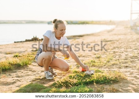 Young female volunteer satisfied with picking up trash, a plastic bottles and coffee cups, clean up beach with a sea. Woman collecting garbage. Environmental ecology pollution concept. Earth Day.