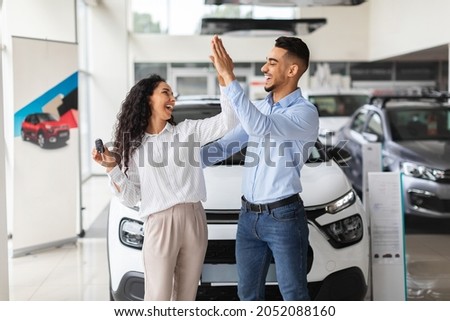 Emotional middle-eastern lovers young man and woman giving high five, standing by nice brand new car, holding key and laughing, buying new auto together at luxury showroom, copy space Royalty-Free Stock Photo #2052088160