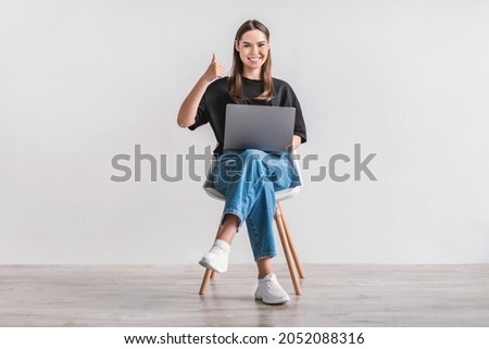 Smiling millennial woman working on laptop, showing thumb up gesture, recommending new website, sitting on chair against white wall. Young lady surfing internet, searching for information online Royalty-Free Stock Photo #2052088316