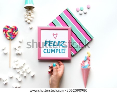 Text Feliz Cumpleanos means Happy Birthday in Spanish. Pink picture frame in hand. Sweets and party decor objects. Candy, snacks, marshmallows. Festive objects on off white background.