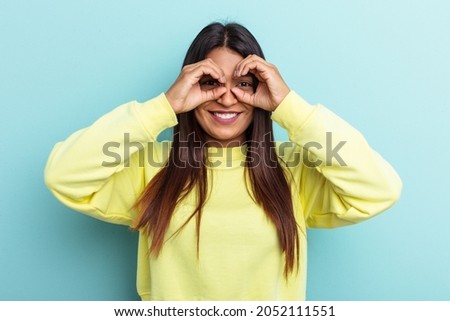 Young Venezuelan woman isolated on blue background showing okay sign over eyes