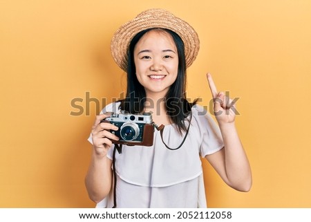 Young chinese girl wearing summer hat holding vintage camera smiling with an idea or question pointing finger with happy face, number one 