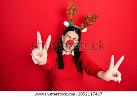Young hispanic woman wearing deer christmas hat and red nose smiling with tongue out showing fingers of both hands doing victory sign. number two. 