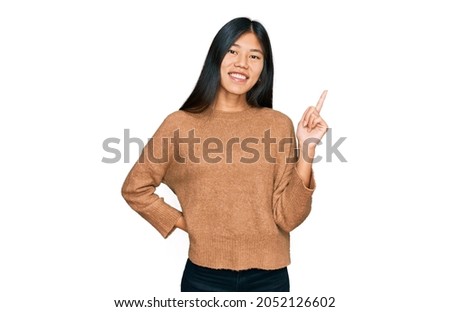 Beautiful young asian woman wearing casual winter sweater with a big smile on face, pointing with hand and finger to the side looking at the camera. 