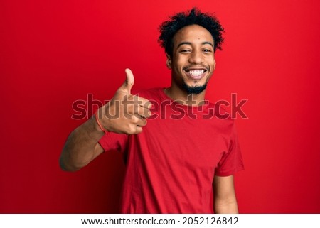 Young african american man with beard wearing casual red t shirt doing happy thumbs up gesture with hand. approving expression looking at the camera showing success. 