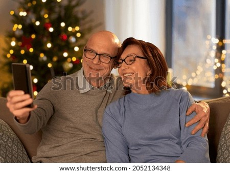 technology, winter holidays and people concept - happy senior couple taking selfie with smartphone at home in evening over christmas tree lights on background
