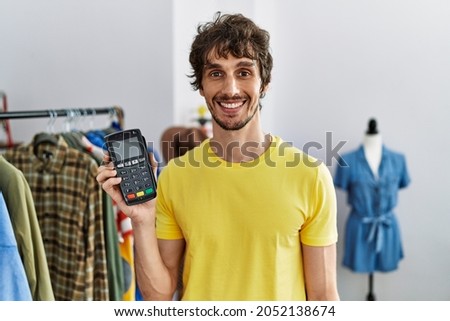 Young hispanic man holding dataphone and credit card at retail shop looking positive and happy standing and smiling with a confident smile showing teeth 