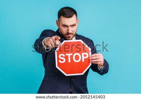 Hey you, access forbidden! Strict bossy man wearing dark official style suit, pointing to camera and holding Stop sign as symbol of prohibition. Indoor studio shot isolated on blue background. Royalty-Free Stock Photo #2052145403