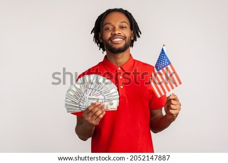 Portrait of smiling satisfied man with dreadlocks wearing red casual style T-shirt, holding american flag and dollars banknotes, celebrating success. Indoor studio shot isolated on gray background.