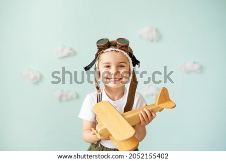 A boy in an aviator helmet and suspenders stands on a blue background with clouds and plays with a wooden plane. Royalty-Free Stock Photo #2052155402