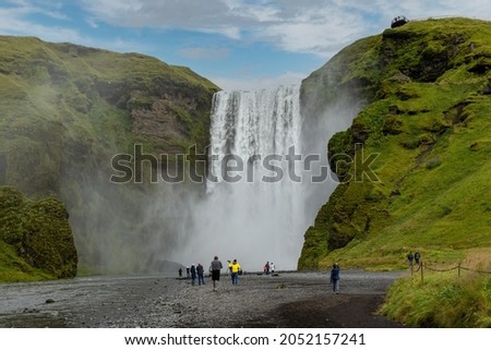 View from the lower part of the Skogafoss waterfall in Iceland with its origin in the volcano glacier Eyjafjallajokull; numerous people walking on river bank to make pictures