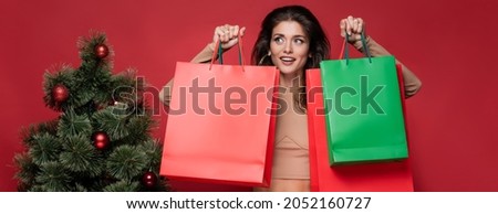 young and joyful woman holding shopping bags near christmas tree isolated on red, banner