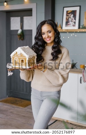 A smiling, happy, attractive young woman holds a Christmas gingerbread house, which she baked herself, in the Scandinavian-style kitchen