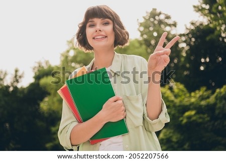 Photo of pretty cute young lady dressed green shirt backpack walking holding books showing v-sign smiling outdoors urban park