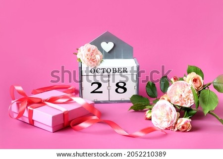 Calendar for October 28: the name of the month in English, the number 28, delicate roses, a packed gift tied with a pink ribbon on a pink background