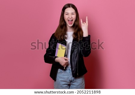 happy cheerful brunette teenager school girl listening to music with headphones depicting heavy metal rock sign wearing a leather jacket and holds books in hands