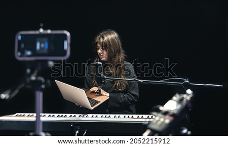 Girl musician with laptop, recording video on smartphone standing on tripod, using professional microphone, blogger or music teacher shooting course in studio, sitting at the piano.