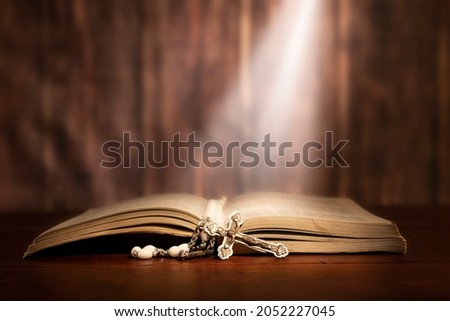 Dramatic image showing a bright light beam shining on an old bible with a rosary laying in front of it.

