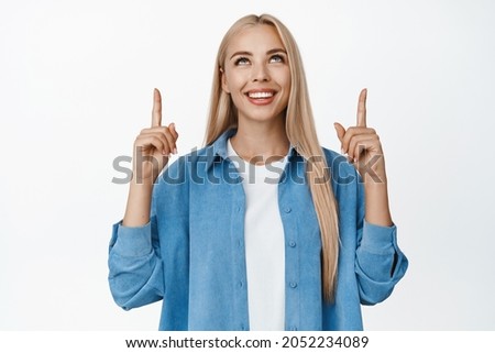 Close up portrait of smiling beautiful lady pointing finger up, looking at advertisement, showing logo, standing over white background