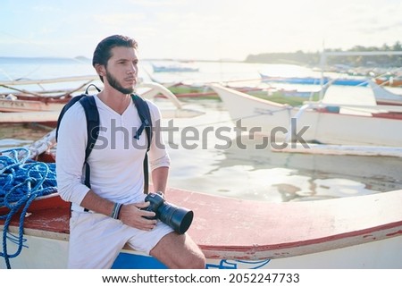 Photography and travel. Young man with rucksack holding camera enjoying beautiful tropical sea view on fishing beach .