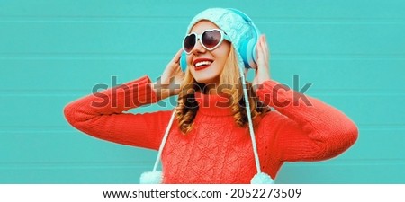 Winter portrait cheerful young woman in wireless headphones listening to music wearing red knitted sweater and white hat with pom pom, heart shaped sunglasses on blue background