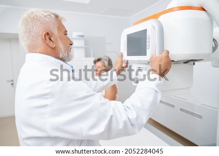 Old grey haired doctor using moving big ultrasound equipment looking at screen, preparing for procedure. Female patient waiting for usg checking of health, sitting in cabinet in modern hospital.