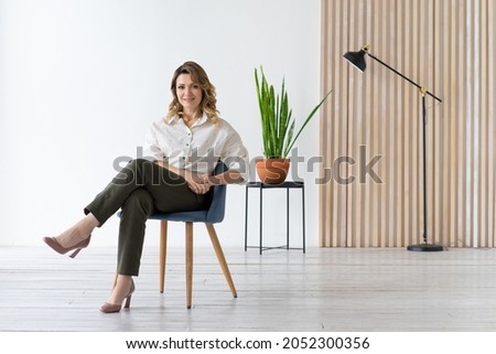 Happy middle-aged woman sits on chair. Minimalistic interior. White background with copy space. Royalty-Free Stock Photo #2052300356