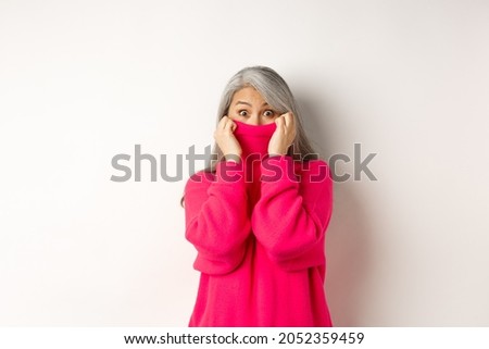 Portrait of funny asian grandmother hiding face in sweater collar, peeking at camera silly, standing over white background