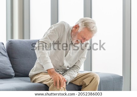 Old man suffering from knee pain sitting sofa in the living room, Elderly man suffering from knee pain while sitting on the sofa Royalty-Free Stock Photo #2052386003