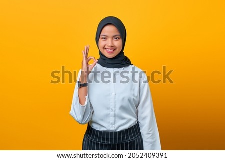 Portrait of happy young Asian woman makes okay gesture on yellow background