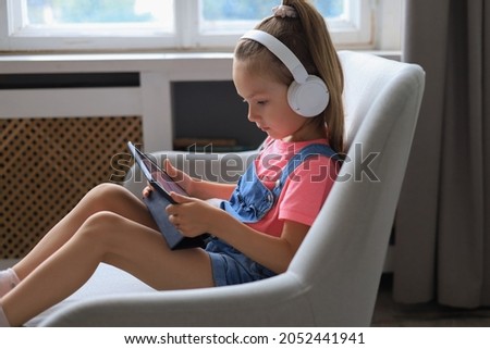 Smiling little girl sitting on sofa playing online games, web surfing information, using funny applications on tablet