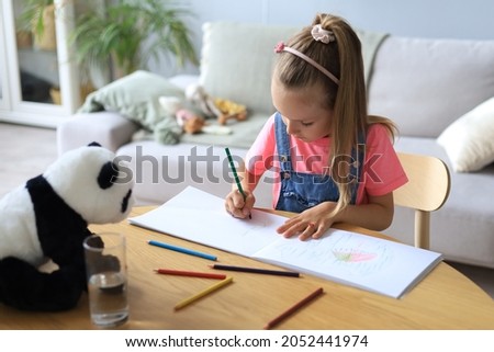 Smilling happy girl sitting in the table with a toy panda bear near to her enjoying creative activity, drawing pencils coloring pictures in albums