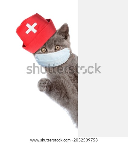 Kitten wearing doctors cap and medical protective mask looks from behind empty banner. Isolated on white background.
